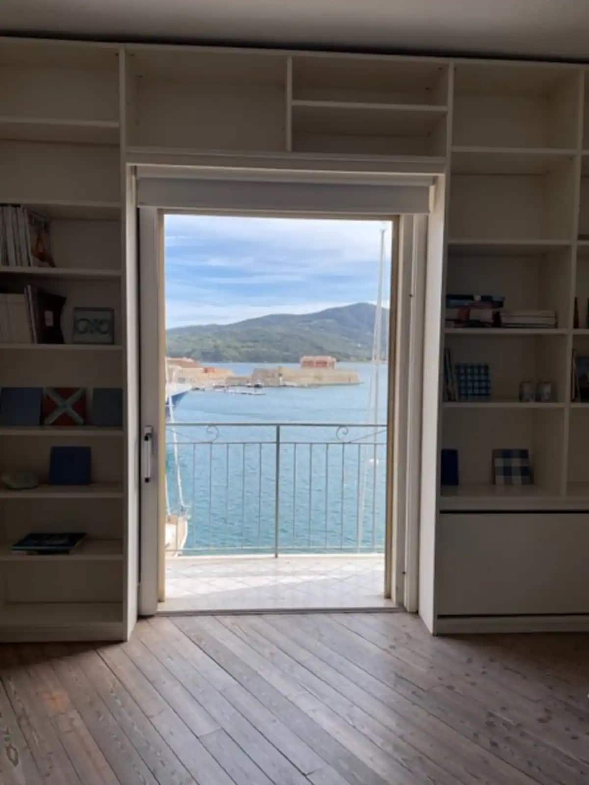 A doorway framed by built-in shelves offers a view of the serene waters of the Darsena Medicea. The soft light illuminates the interior, while distant hills and boats can be seen through the open door, creating a peaceful coastal ambiance.