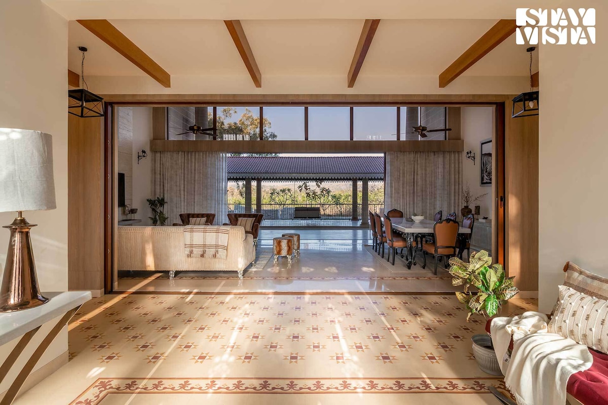 A spacious living area is showcased with warm wooden beams and large glass doors that invite natural light. Comfortable seating is arranged alongside a dining table, creating an open flow towards the outdoor area, visible through the expansive windows.