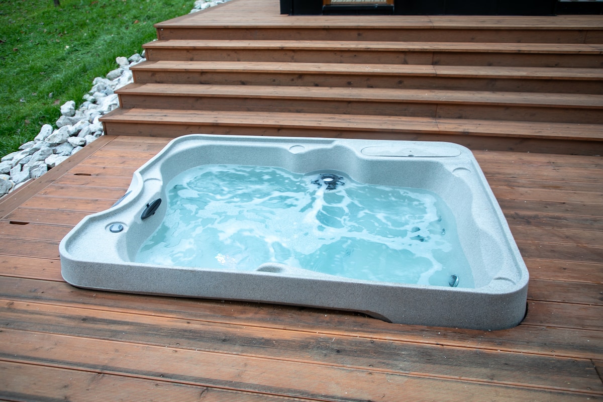 A private hot tub with a textured gray surface is positioned on a wooden deck. The water appears clear and inviting, contrasting with the surrounding greenery and the natural wood finish of the elevation.