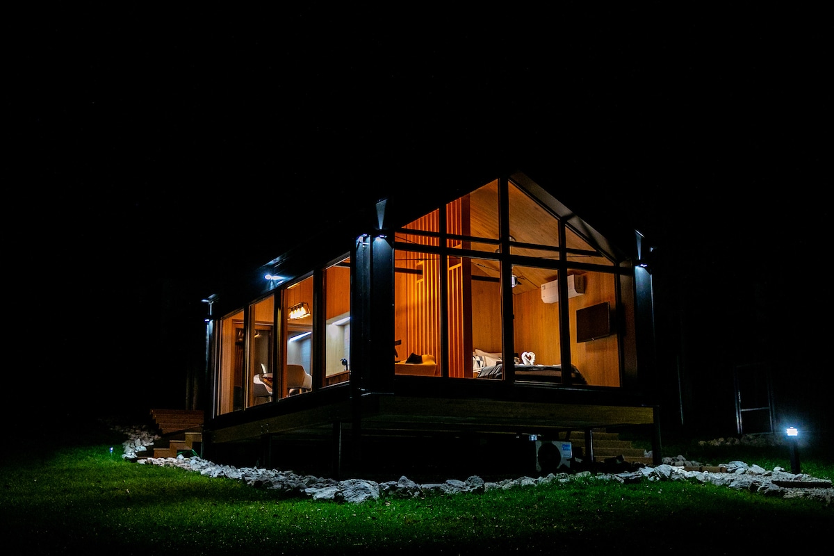 The modern guesthouse, illuminated at night, features large glass windows that provide a view of the cozy interior. The surrounding yard is landscaped with rocks, enhancing the peaceful ambiance of the wooded setting.