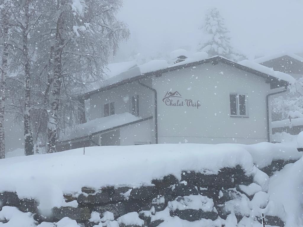 The exterior of Das Chalet Weiß is seen in a winter setting, surrounded by falling snowflakes. A layer of snow covers the roof and stone wall. The chalet's name is displayed on the façade, with windows partially visible behind the snow-covered surroundings.