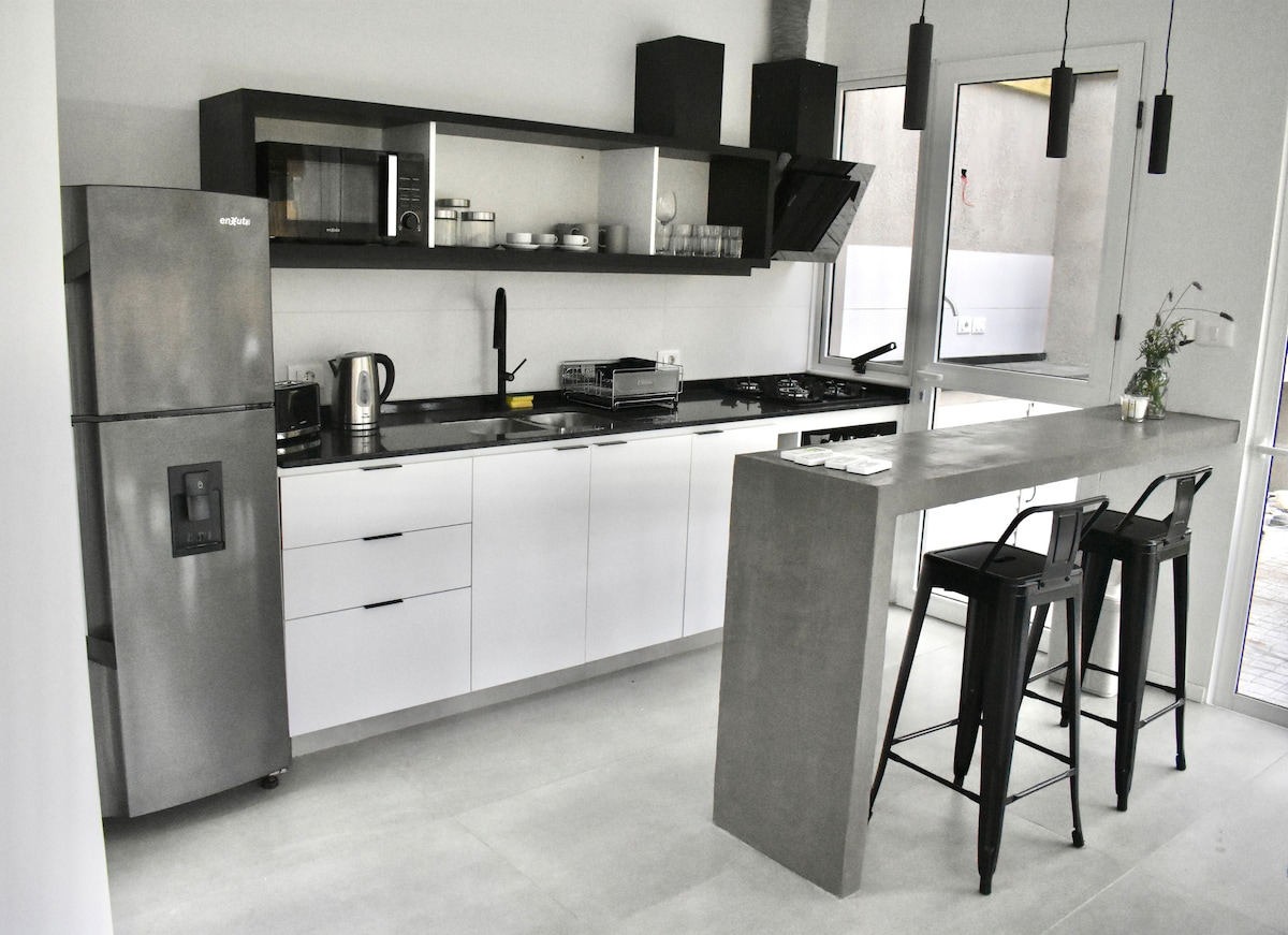 A modern kitchen space is shown, featuring sleek black and white cabinetry. A refrigerator and essential appliances are displayed on the counter, which has a minimalist concrete bar area with high stools. Large windows allow natural light to enter the open layout.