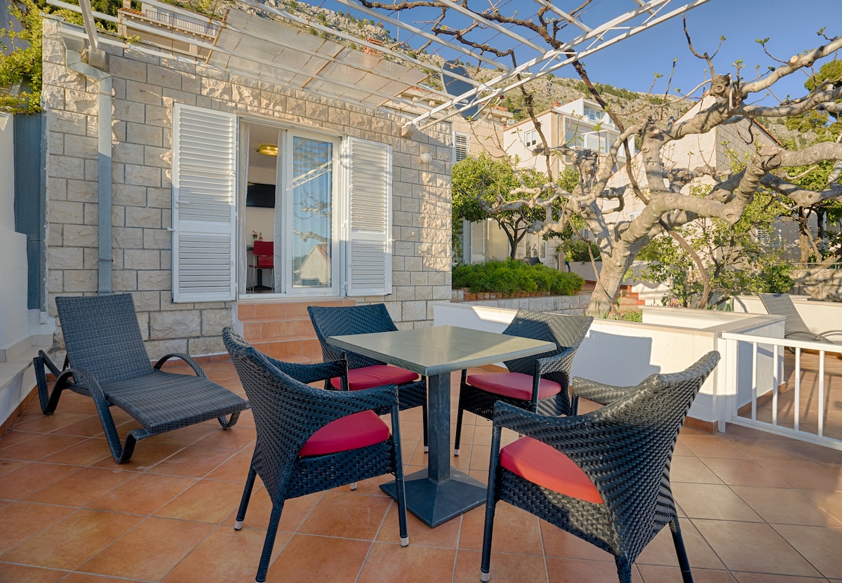 An outdoor terrace is furnished with a square table surrounded by four wicker chairs featuring red cushions. Two lounge chairs are positioned nearby, offering relaxation options. The stone structure showcases glass doors that lead into the interior space, complemented by greenery in the surrounding area.