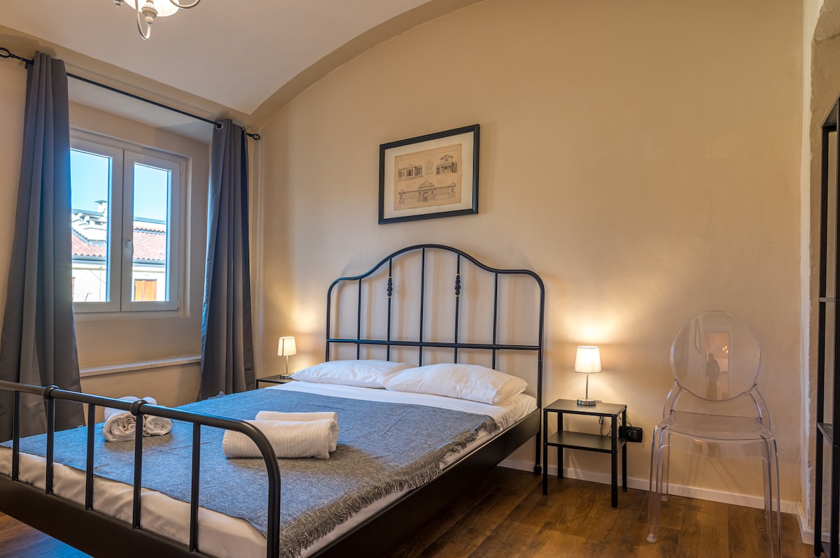 A well-appointed bedroom is depicted with a spacious bed featuring a sleek black metal frame. Soft bedding is neatly arranged with towels placed at the foot. Natural light enters through window drapes, and a small bedside table holds a lamp on each side, accompanied by a transparent chair.