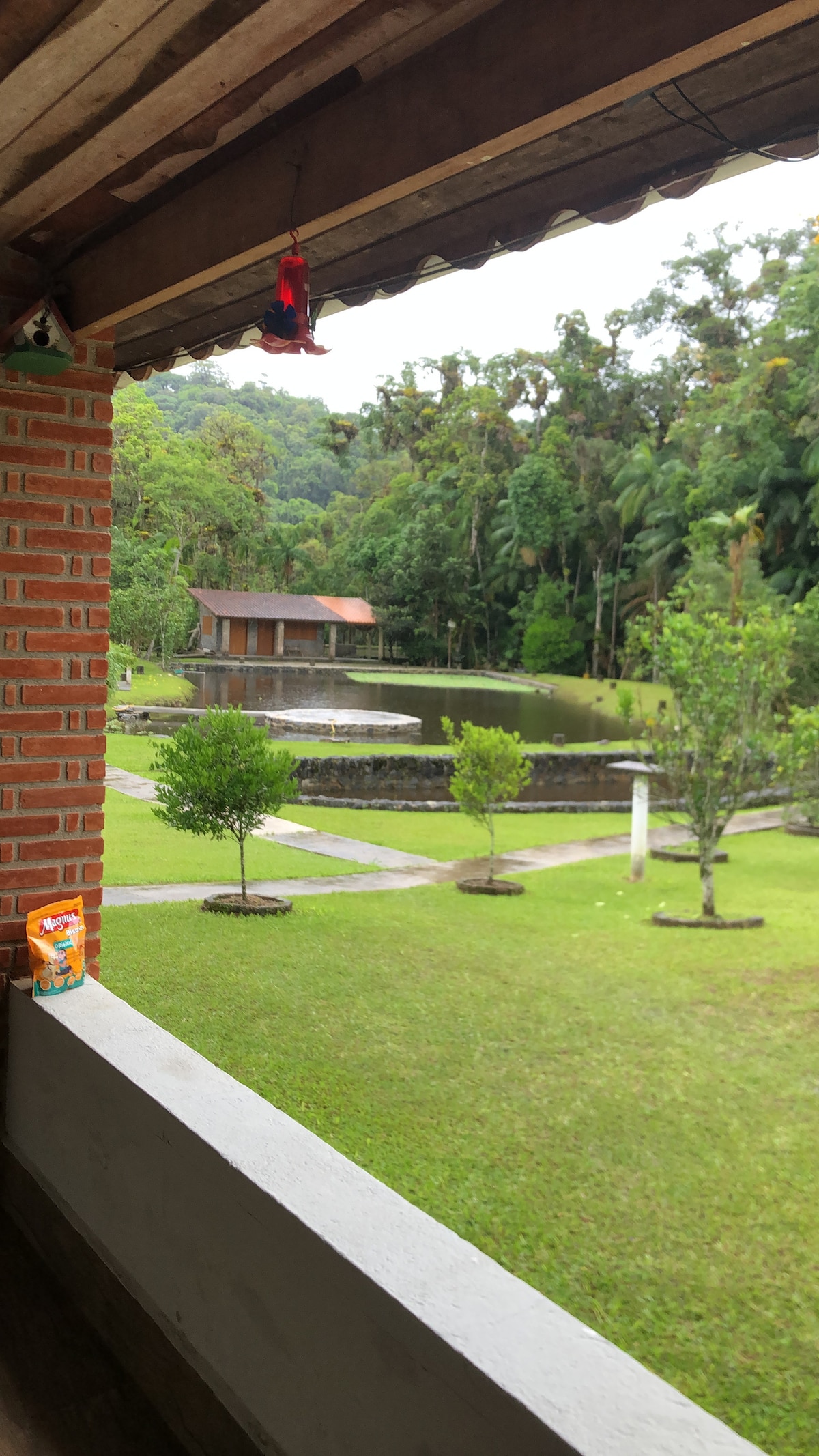 A view from a covered porch showcases a serene lake bordered by lush greenery. A small wooden structure with a red roof is visible on the opposite side of the water. Neatly trimmed grass and young trees frame the landscape, contributing to a peaceful atmosphere.