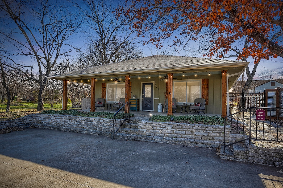 The cottage exterior showcases a welcoming porch with seating arranged for relaxation. The stone foundation contrasts with the wooden accents and the light fixtures overhead. Surrounding trees provide a natural ambiance, while a spacious driveway leads up to the entrance.