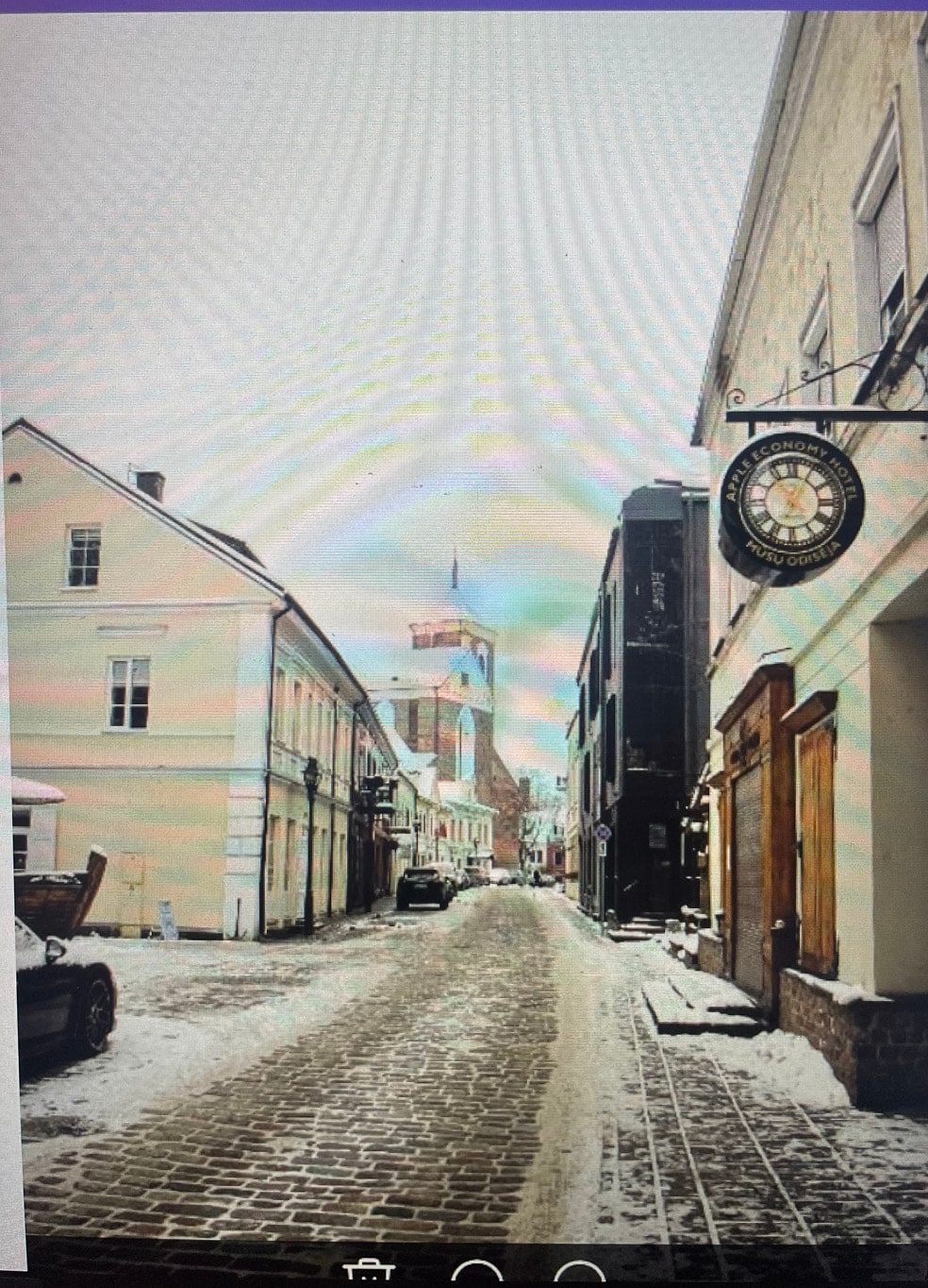 A snow-covered street is lined with historical buildings, showcasing a mix of light and dark façades. A clock tower is visible in the distance, surrounded by gently falling snow. The cobblestone pathway reflects a serene winter scene, with vehicles parked along the side.