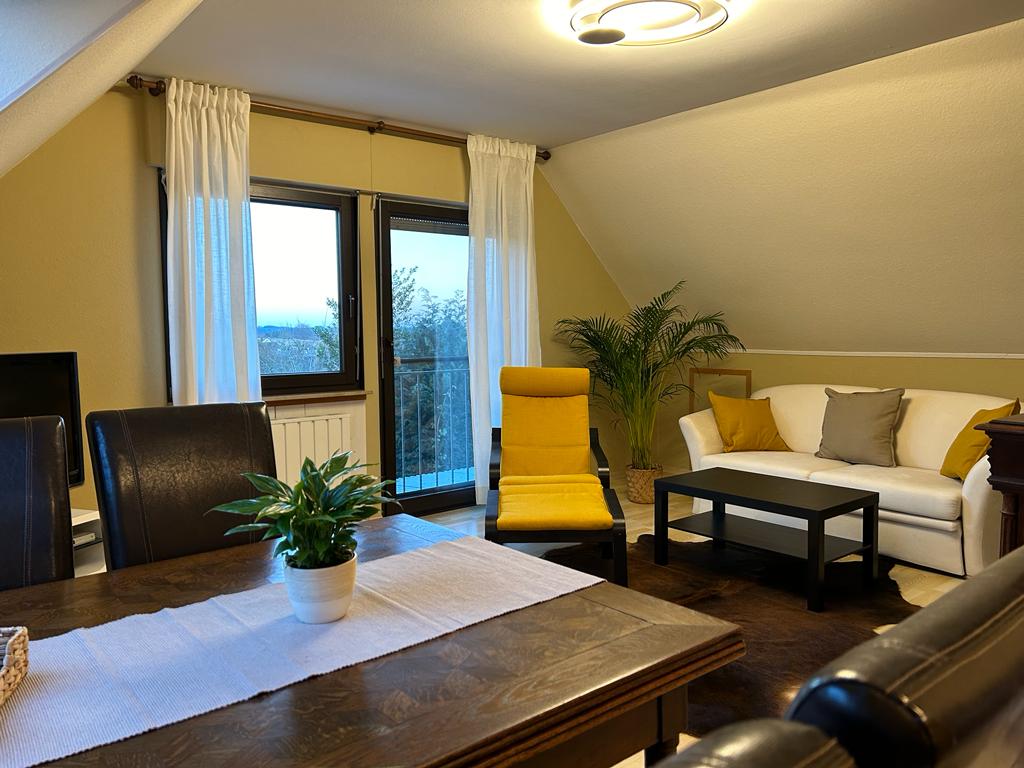 A well-lit living area features a spacious white sofa, a yellow accent chair, and a dark coffee table. Large windows invite natural light and offer views outside, while a small indoor plant adds a touch of greenery. The dining table is set for four.