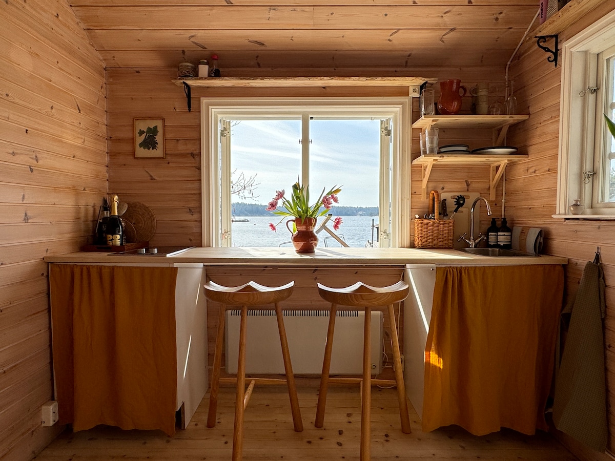 A cozy kitchen space features a wooden countertop with two stools. Sunlight flows in through a wide window, offering a view of the water. A vase of fresh flowers sits on the counter, surrounded by wooden shelves displaying kitchen items.