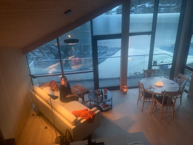 A modern living area features large glass walls offering views of the serene outdoor landscape. A cozy sofa is complemented by a warm throw, and a coffee table is arranged with drinks. A dining table with chairs is visible in the background, creating an inviting atmosphere.