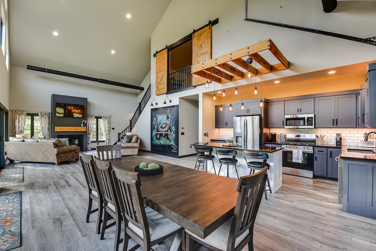 An open-concept living area features high ceilings and large windows allowing natural light. A spacious dining table is surrounded by wooden chairs. The kitchen is equipped with modern appliances, and a cozy seating area includes a comfortable sofa, creating a welcoming gathering space.