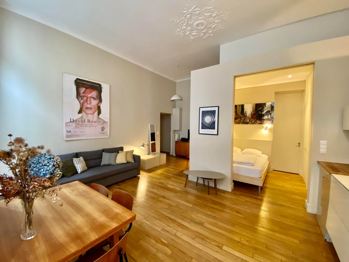A stylish living area features a comfortable gray sofa and a wooden dining table surrounded by chairs. Light wood flooring creates a warm atmosphere. An open doorway leads to a sleeping area with a spacious bed, while art pieces adorn the walls.