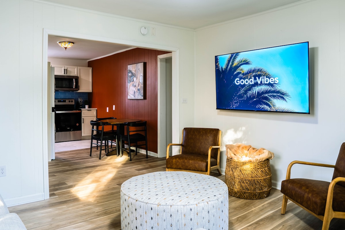 A comfortable living area features two armchairs and a round coffee table. A large wall-mounted TV displays an inviting message. Natural light enhances the space, highlighting the warm wood flooring and the adjacent dining area with a black table and four chairs.