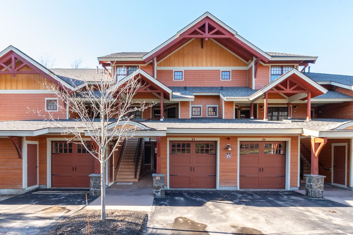 The exterior of a multi-story condo building is presented, featuring a combination of wood and stone finishes. Garages with brown doors are visible at the ground level, along with a pathway leading to the main entrance, framed by a landscaped area.