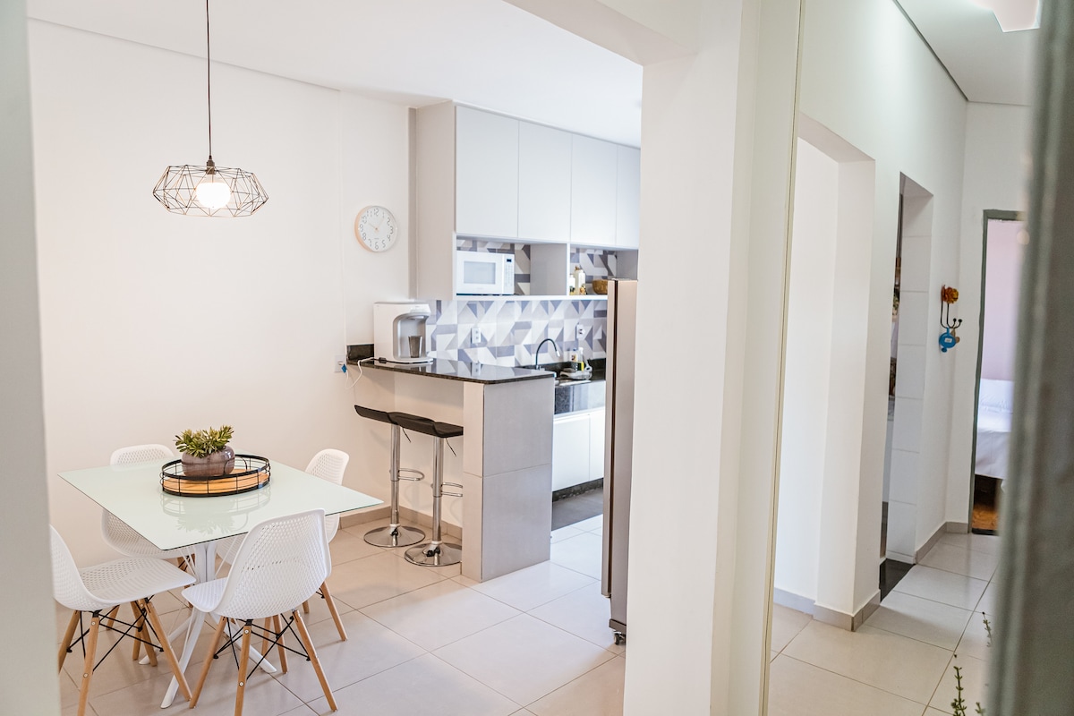 A modern kitchen and dining area is visible, featuring a sleek white table surrounded by plastic chairs. A minimalist pendant light hangs above the table, while appliances, including a microwave and refrigerator, line the expansive countertop. Natural light brightens the open space.