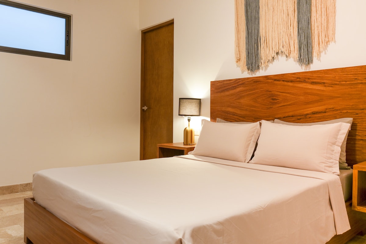 A well-furnished sleeping area features a spacious bed dressed in neutral linens. Wooden bedside tables with lamps complement the modern decor, while a textured wall hanging adds visual interest. Natural light enters through a small window, enhancing the calm atmosphere.