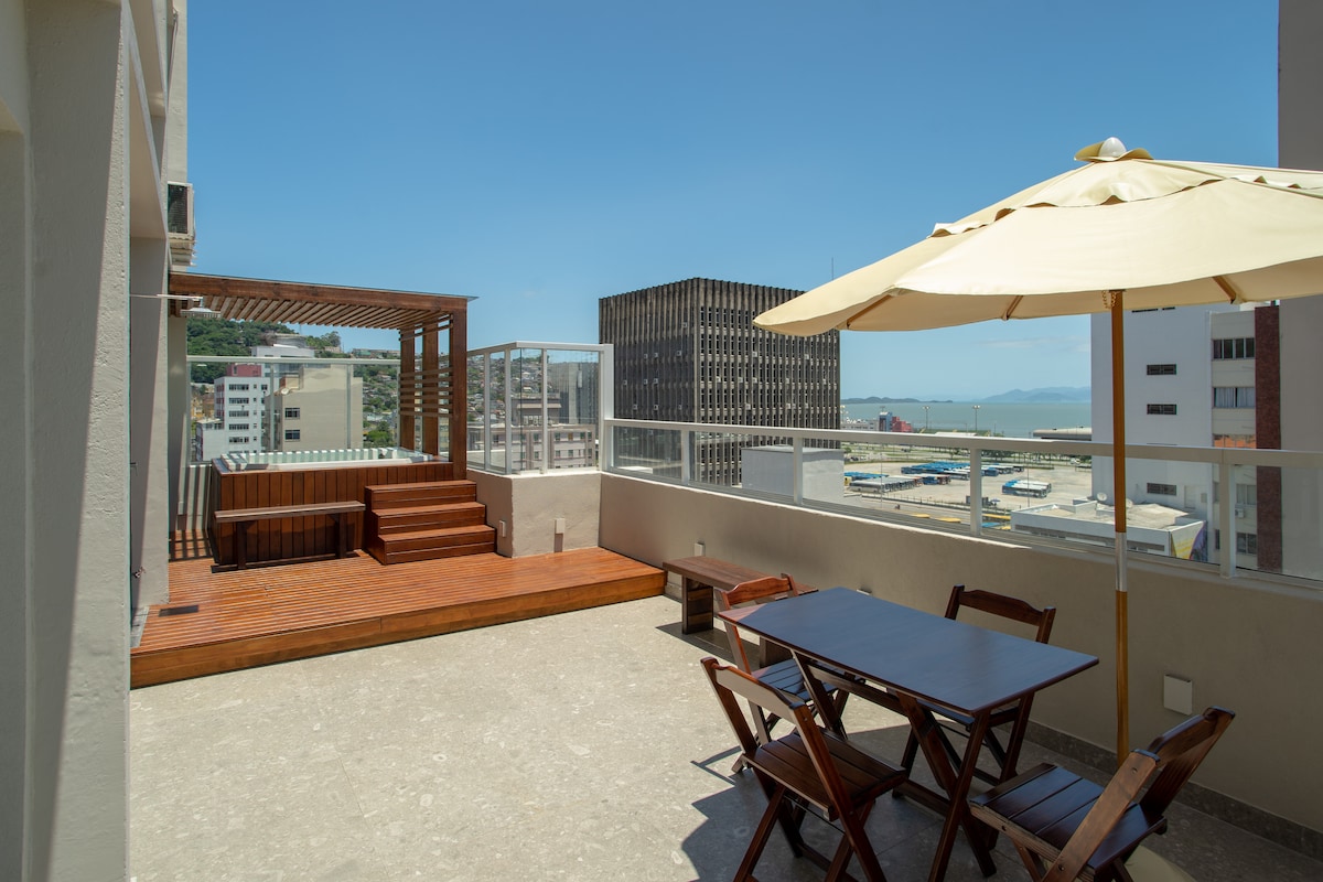 An outdoor terrace features a wooden deck and a pergola, providing a shaded area with a sun umbrella. A dining table and chairs are positioned on one side, with scenic views of the city and distant water visible in the background.