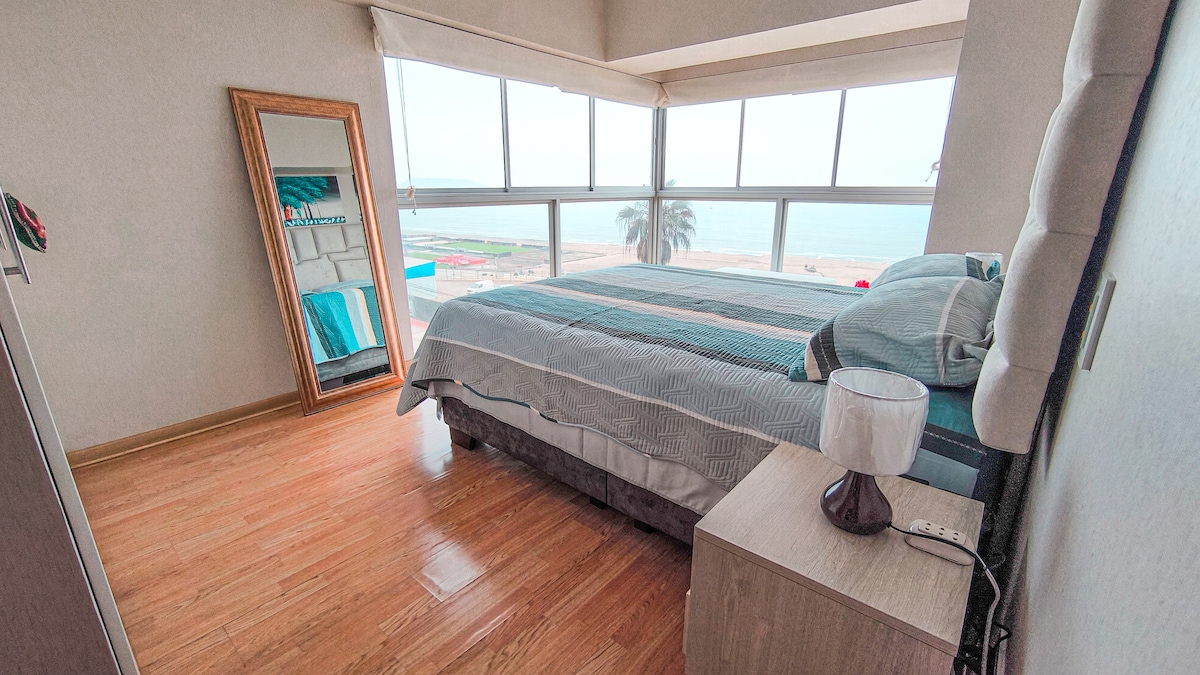 A cozy bedroom is highlighted by large windows offering views of the ocean. A spacious bed with soothing striped bedding is positioned beside a small nightstand featuring a lamp. A mirror reflects the interior, contributing to the bright and airy atmosphere.
