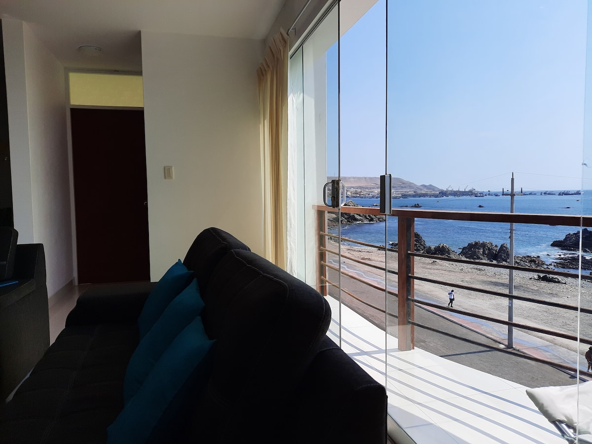 A comfortable seating area is positioned near large glass windows, offering a clear view of the ocean and shoreline. Soft cushions in shades of blue add color to the dark furniture. Natural light brightens the space, complemented by serene coastal scenery.