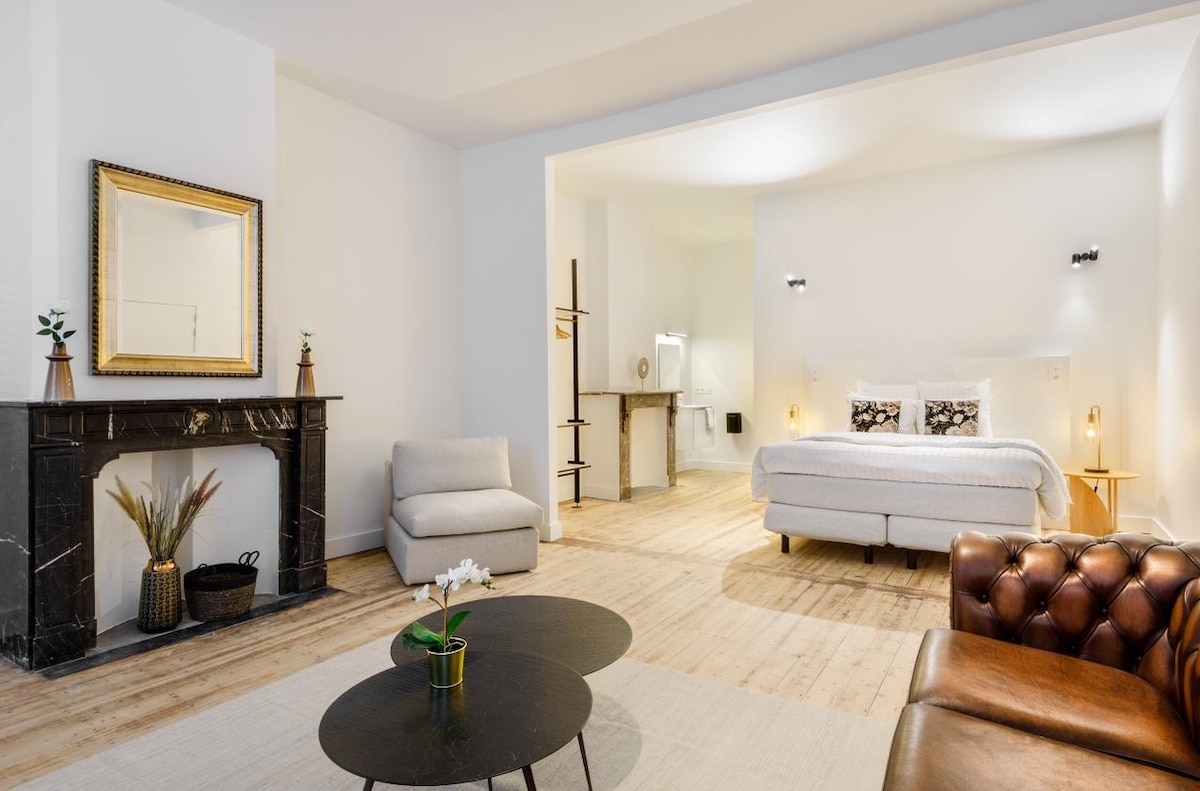 A spacious room features a queen-size bed with quality linens, complemented by a cozy seating area. A dark marble fireplace adds elegance, while a large mirror reflects light across the hardwood floor. Soft lighting enhances the serene ambiance of the suite.