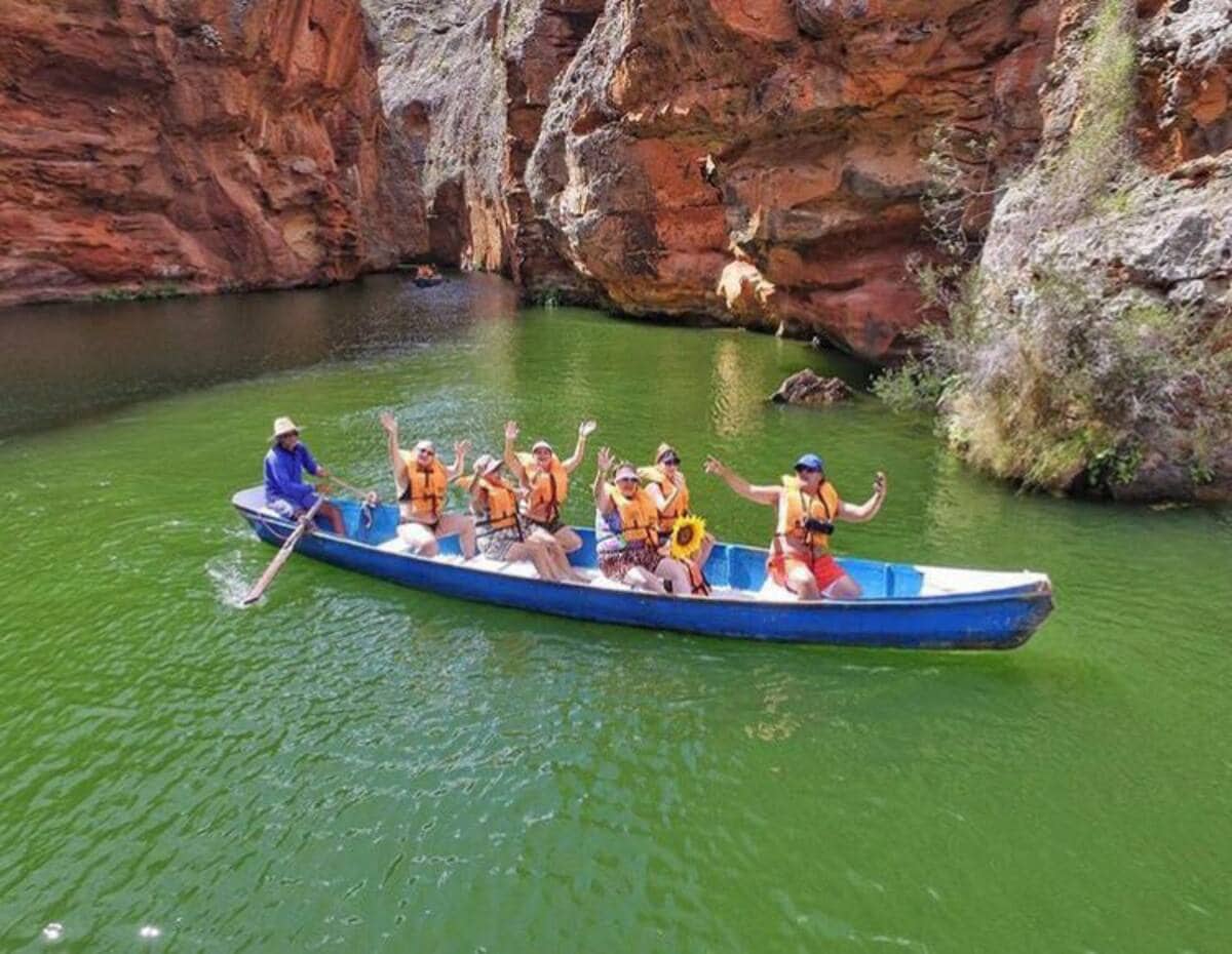 A group of six individuals in orange life jackets is seated in an elongated blue boat, navigating through calm green waters surrounded by towering reddish cliffs. The joyful expressions and raised arms of the participants convey a sense of camaraderie amidst the serene natural setting.
