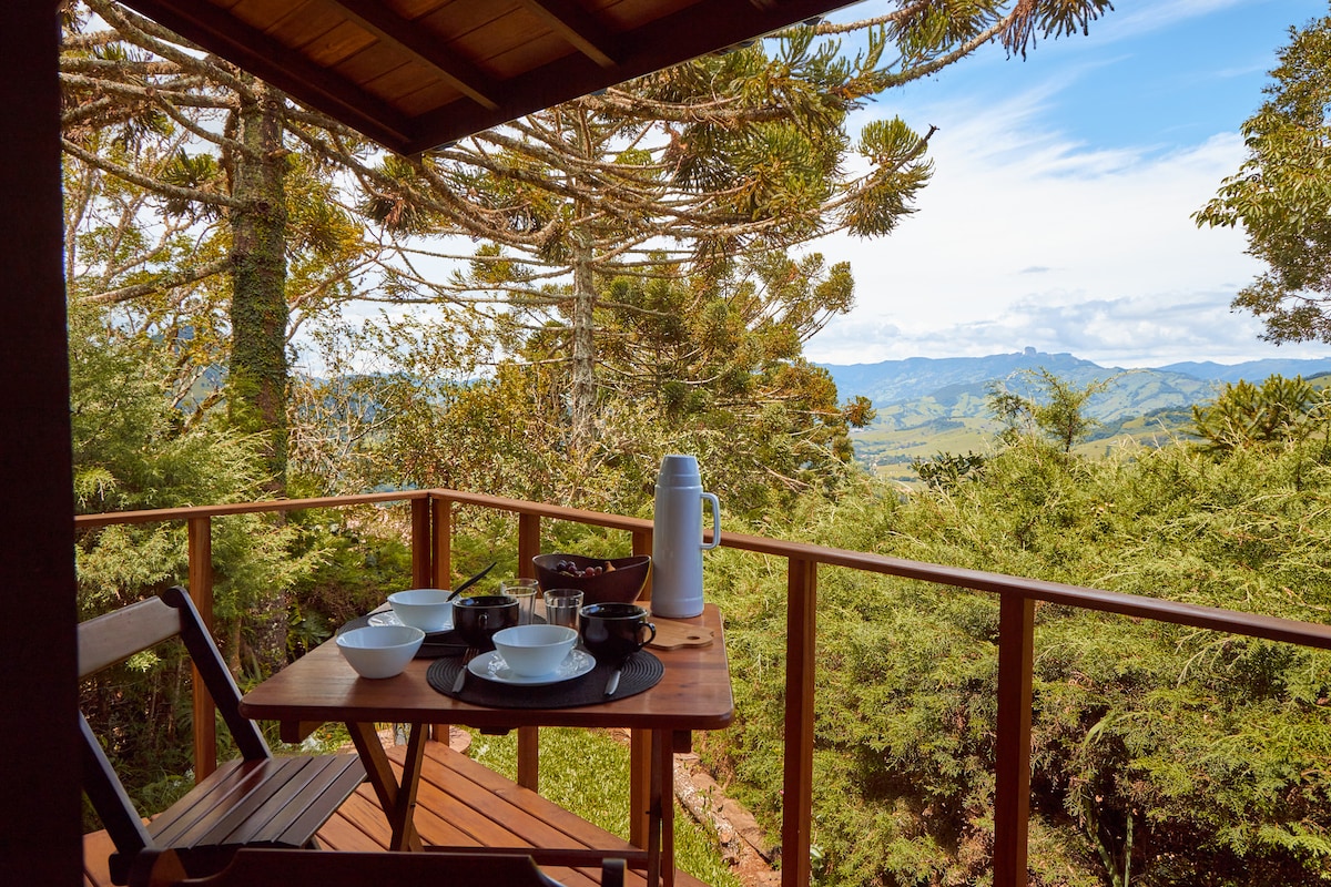 A wooden balcony features a cozy dining setup with a small table and two chairs. Bowls and tableware are arranged neatly, accompanied by a thermos. Lush greenery and a scenic landscape are visible in the background, enhancing the tranquil atmosphere.