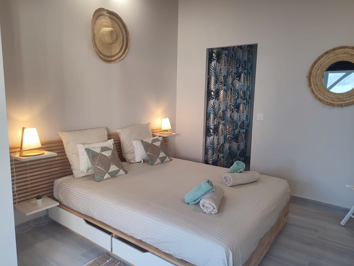 A cozy sleeping area is presented, featuring a spacious bed with neutral-colored linens. Two bedside lamps provide soft lighting. Decorative pillows add texture, while a small shelf holds a bedside clock. A leaf-patterned curtain complements the room's design.