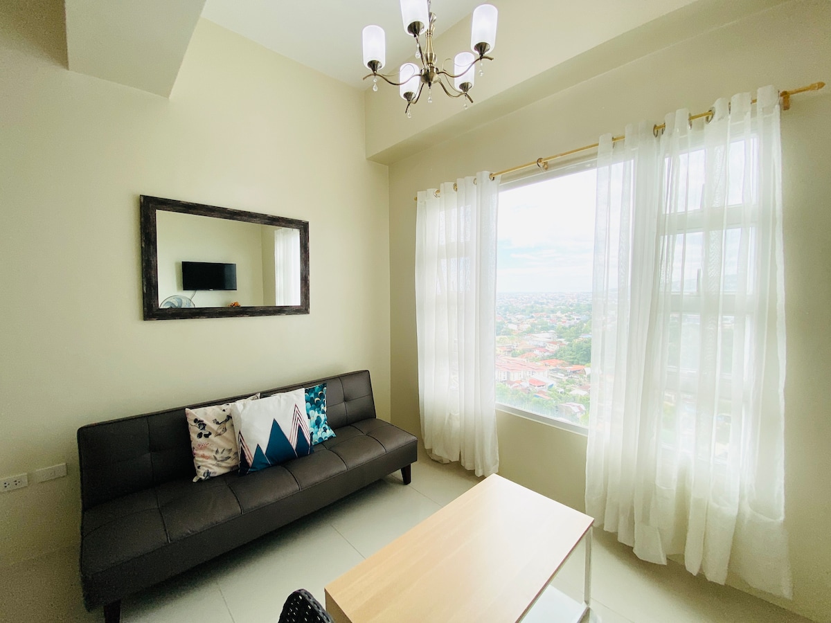 A well-lit living area features a dark sofa adorned with decorative pillows, paired with a modern wooden table. Large windows, framed by sheer white curtains, offer panoramic views of the cityscape, while a mirror reflects light and enhances the sense of space.