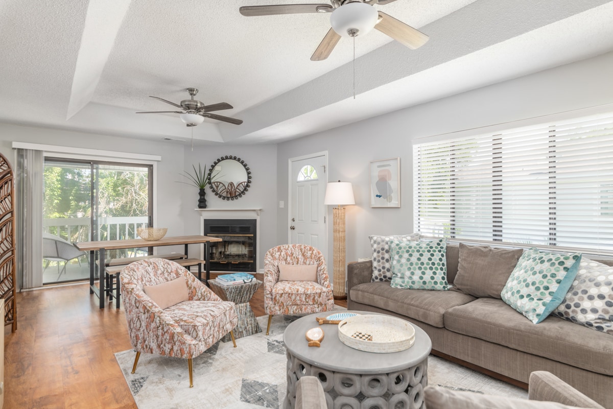 A bright living room features comfortable seating arrangements with floral accent chairs and a sectional sofa. Natural light filters through large windows, highlighting a coffee table and a decorative rug. A ceiling fan and wall art add to the welcoming atmosphere.