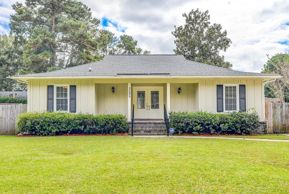 A single-story home features a light yellow exterior complemented by black shutters. The well-maintained lawn is surrounded by neat landscaping, showcasing low shrubs. A set of steps leads to a central entrance, flanked by two windows on each side, creating a welcoming facade.
