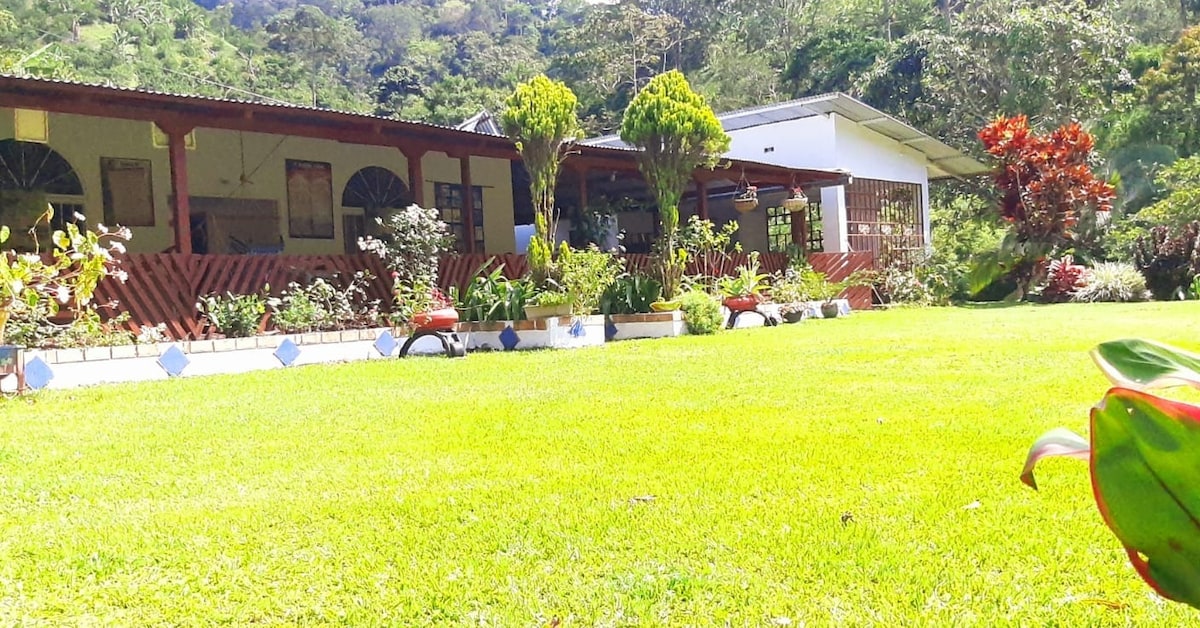 A vast green lawn is surrounded by colorful flower beds and shrubs. Two buildings, one featuring a rustic wooden facade and the other a more modern design, are positioned behind the lush greenery. The serene landscape is dotted with trees, enhancing the tranquil atmosphere.