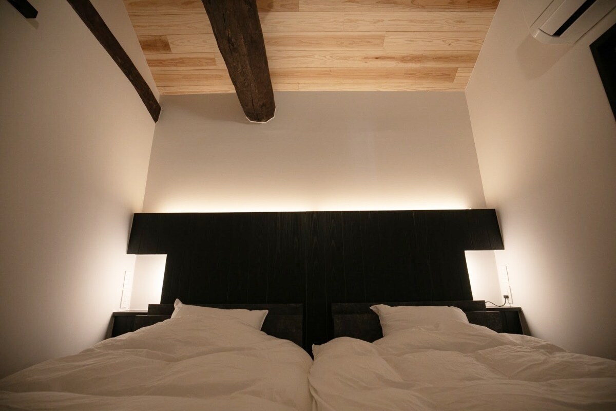 The sleeping area features two spacious beds, each dressed in white linens. Soft, ambient lighting illuminates the headboard, while exposed wooden beams add a rustic touch to the ceiling.