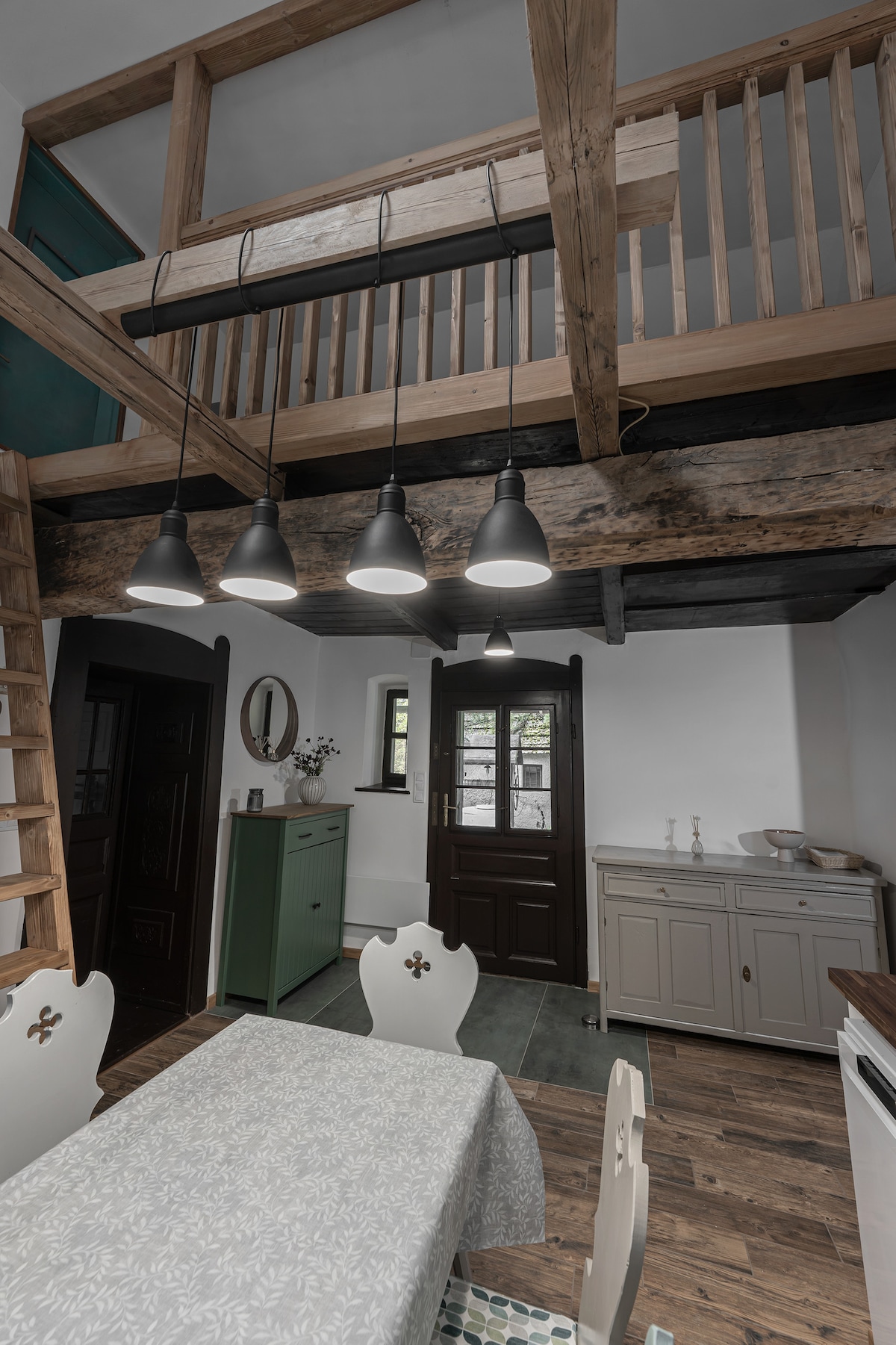 An inviting dining area features a light-colored table with a floral tablecloth, surrounded by white chairs. Above, pendant lights hang from wooden beams, illuminating the space. A staircase leads to a loft area, while a green cabinet and a sideboard add functional decor.