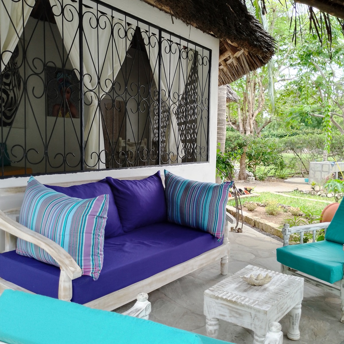 An outdoor seating area features a white wooden couch adorned with vibrant purple cushions, complemented by blue armchairs. A low coffee table sits in the center, surrounded by lush greenery and a tranquil garden view visible through decorative ironwork windows.