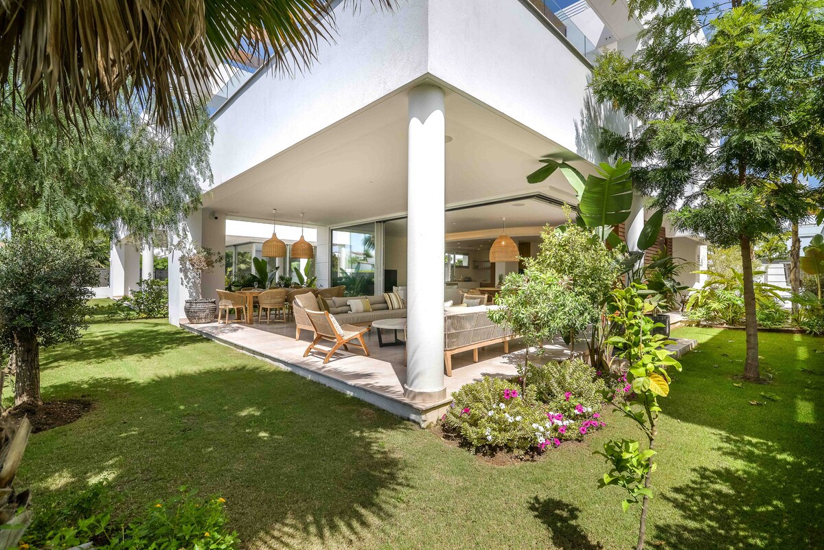 The villa's exterior is framed by lush greenery and colorful flowers. A spacious patio with comfortable seating is visible, complemented by modern light fixtures. The clean lines of the architecture highlight an open feel, creating a seamless blend between indoor and outdoor living areas.