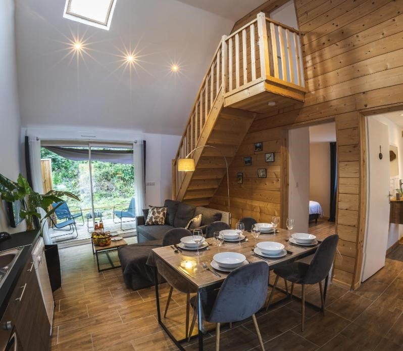 An inviting living space features a dining area with a table set for six and a cozy couch facing a large glass door that opens to a private outdoor area. A wooden staircase leads to additional sleeping accommodations. Natural light illuminates the warm, wooden accents.