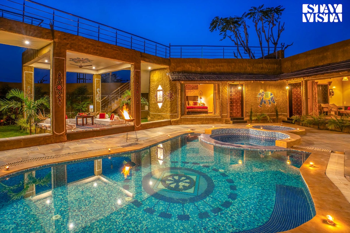 An outdoor swimming pool, featuring intricate mosaic tiles, is illuminated by soft lighting. The pool area is surrounded by a spacious patio, with seating arrangements and elegant decor visible in the background. The tranquil setting merges modern amenities with the natural beauty of the surrounding landscape.