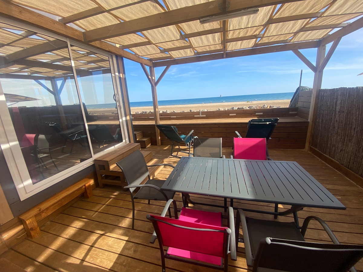 A sheltered outdoor terrace is depicted, featuring a wooden deck and a table surrounded by chairs. In the background, the beach and sea are visible, framed by a clear sky. Sun loungers provide additional seating for relaxation.