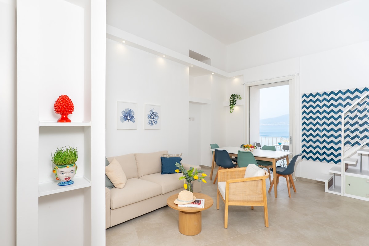 A bright living area is presented, featuring a light-colored sofa and a circular wooden coffee table. A dining space with a table and colorful chairs is visible, along with a large window that offers views of the sea. Wall decor and plants add a fresh touch.