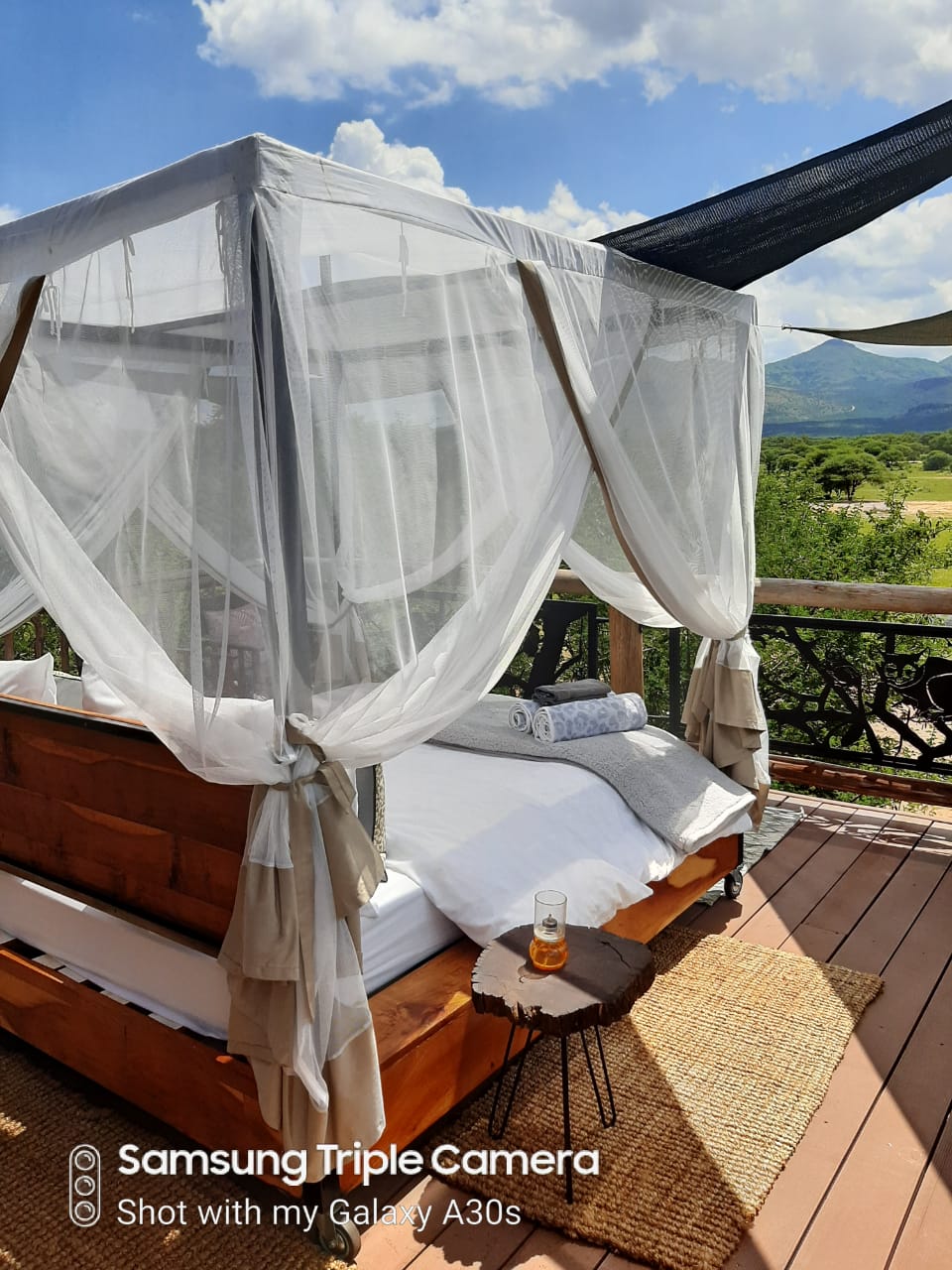 An elevated wooden bed is surrounded by sheer white drapes, allowing for a clear view of the landscape. Soft pillows are arranged at the head of the bed, and a towel is neatly placed nearby. A glass rests on a side table, reflecting natural light.