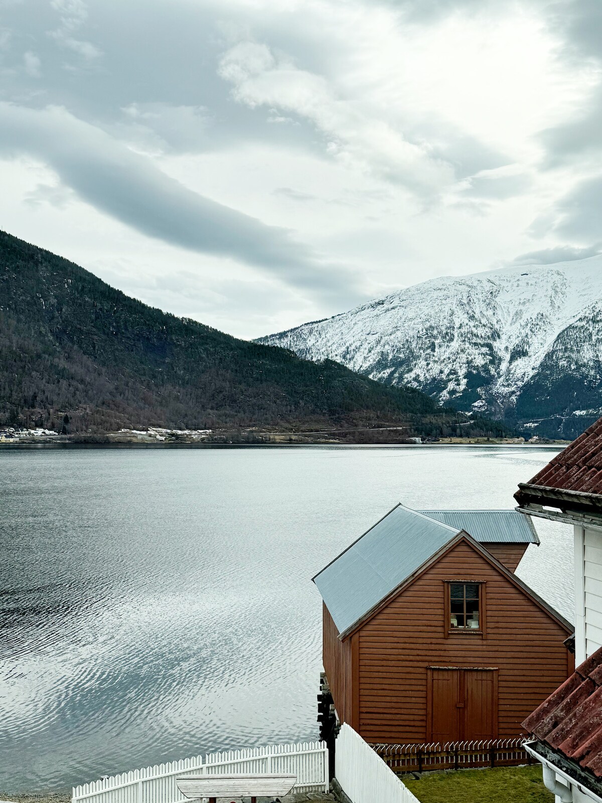 A view of a brown wooden building situated by the fjord, with mountains rising majestically in the background. The calm water reflects the sky and surrounding landscape, while the peaked roof of the building adds architectural interest against the serene setting.