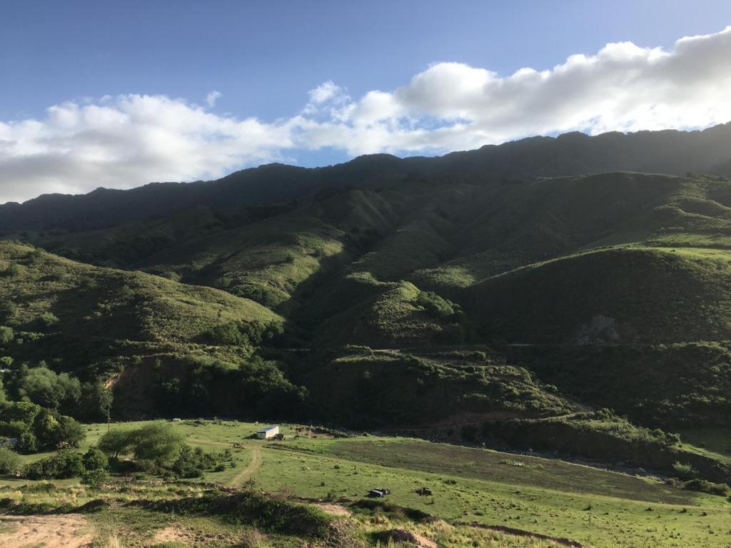 A panoramic view of rolling green hills stretches across the landscape, highlighted by gentle slopes and rugged mountain peaks under a partly cloudy sky. The natural terrain showcases a vibrant mix of foliage, creating a serene backdrop with hints of distant structures visible in the valley.