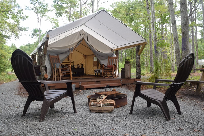 A spacious canvas glamping tent is set among trees, featuring a wooden deck with seating and a fire pit in front. Two dark chairs are positioned nearby, inviting relaxation. Gravel paths surround the area, emphasizing the tent's placement in a natural wooded setting.