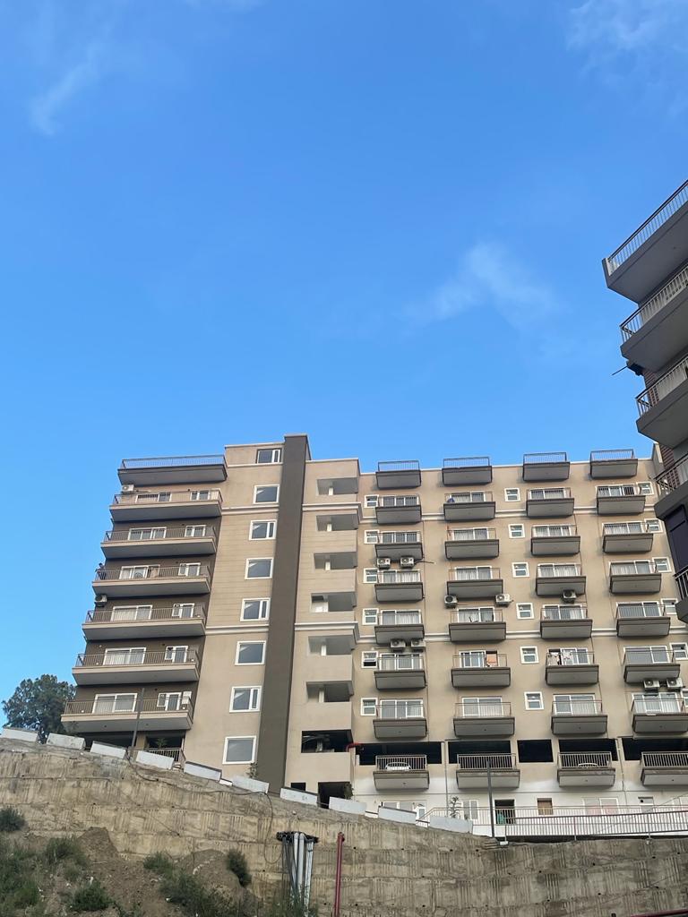 A modern apartment building is depicted against a clear blue sky. The structure features multiple floors, with various balconies and air conditioning units visible. The lower portion of the building is accented by a concrete wall, creating a contrasting foreground.