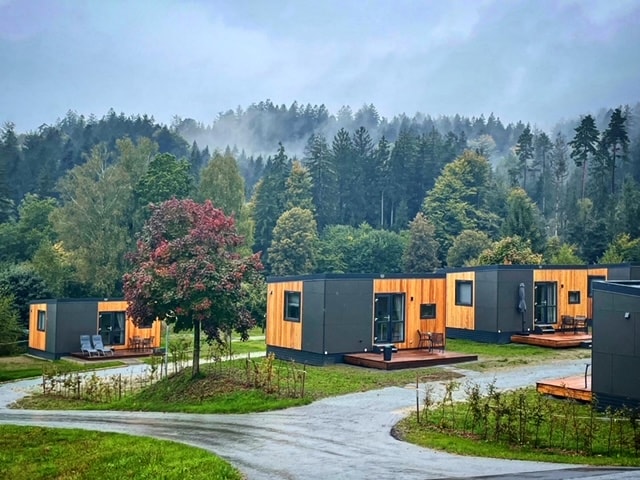 Several modern tiny houses are nestled among greenery, featuring wood accents and large windows. They are situated around a winding driveway, with trees and misty hills in the background, creating a serene environment.