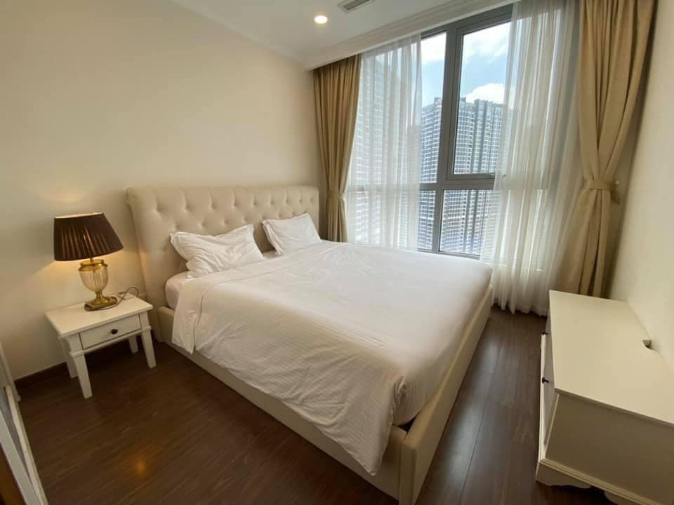 A bright bedroom features a spacious bed with a light-colored duvet. Tall windows dressed in sheer and solid curtains allow natural light to fill the room. A bedside table with a lamp is positioned beside the bed, enhancing the room's warm aesthetic.