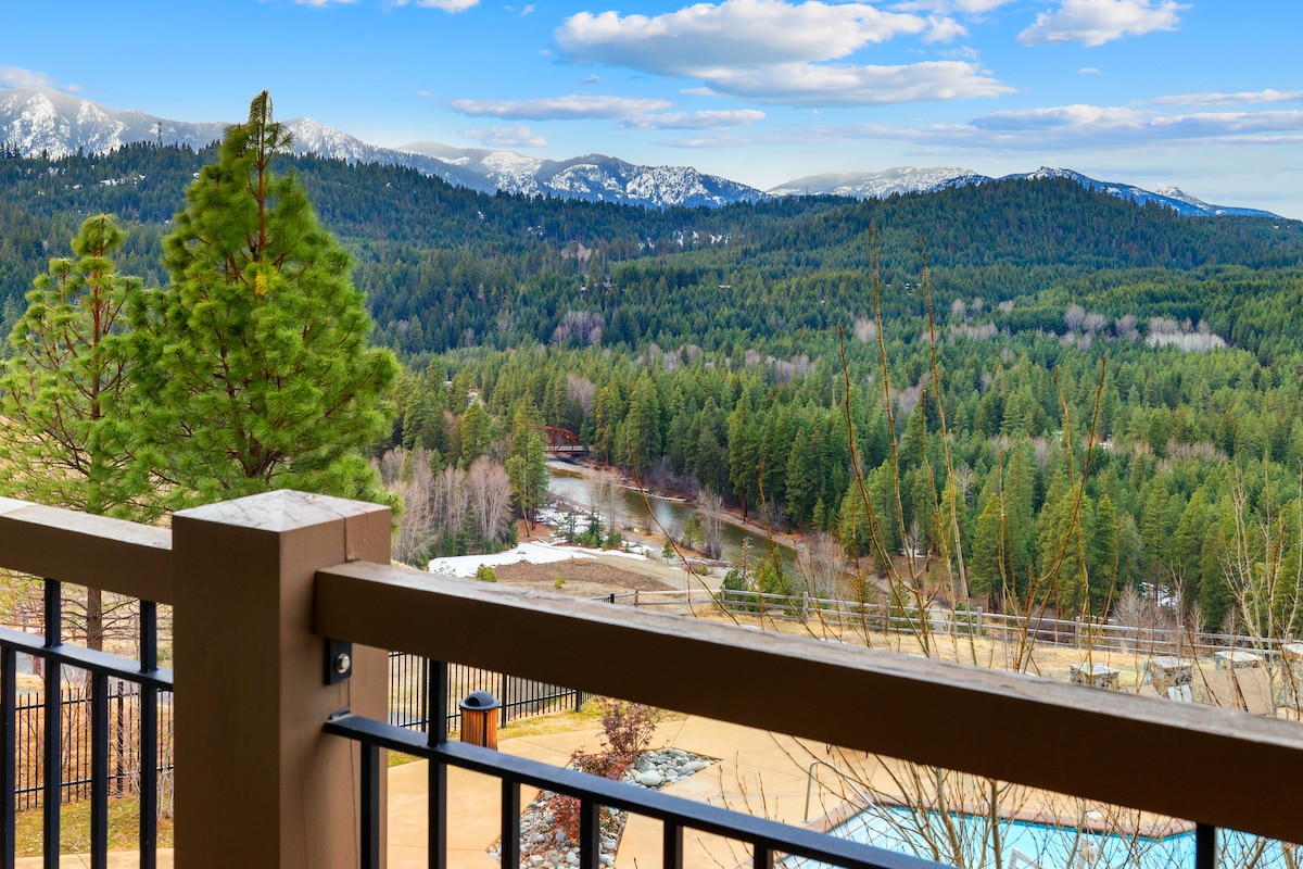 A scenic view from the balcony showcases a vast expanse of lush green trees and rolling mountains under a clear blue sky. The peaceful river can be seen winding through the valley, with hints of snow visible in the landscape.