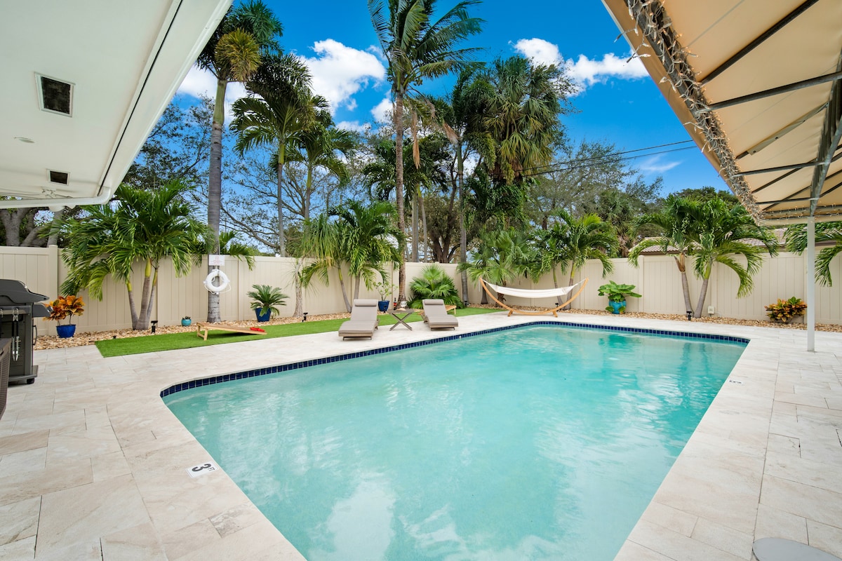 A serene outdoor space features a heated pool surrounded by lush palm trees. Two lounge chairs sit by the water’s edge, while a hammock is positioned nearby for relaxation. The patio is bordered by well-maintained landscaping, providing a tranquil atmosphere for leisure.