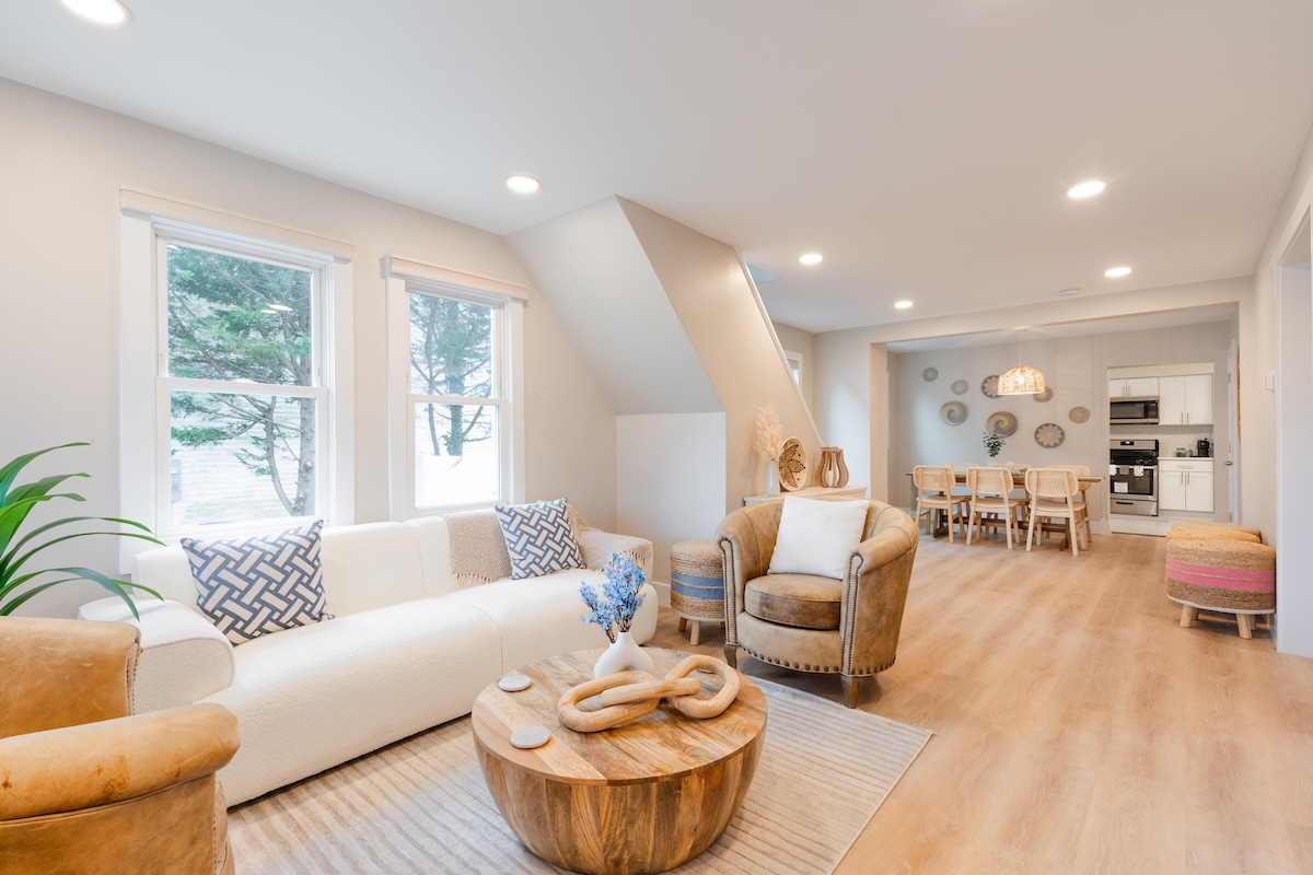 A bright living area is highlighted by natural light from two windows. A white sofa and a cozy armchair are positioned around a round wooden coffee table. A dining area with a wooden table and chairs is visible in the background, complemented by unique wall decor.