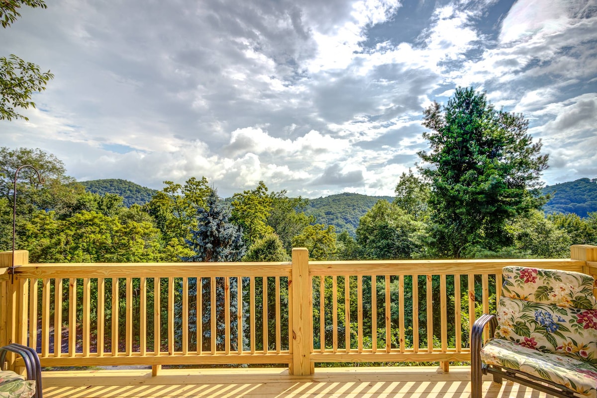 A wooden deck features comfortable seating surrounded by lush greenery. The expansive view includes rolling mountains under a partly cloudy sky, providing a serene backdrop. The railing is crafted from timber, adding to the natural aesthetic of the outdoor space.